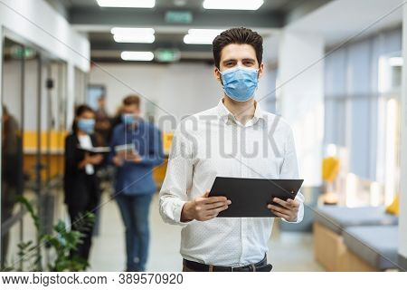 Portrait Of A Businessman At The Office In A Mask. A Group Of Collegues Discuss Business Matters At 