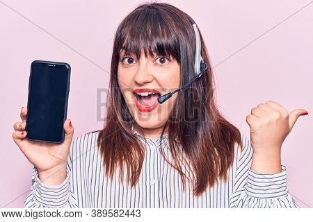 Young plus size woman wearing call center agent headset holding smartphone pointing thumb up to the side smiling happy with open mouth 