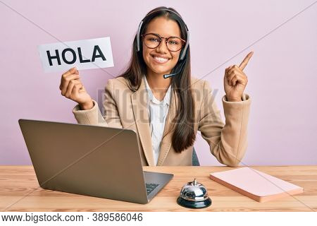 Beautiful hispanic woman wearing operator headset holding hola greenting smiling happy pointing with hand and finger to the side 