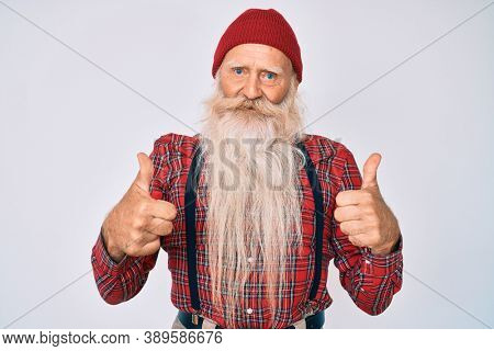 Old senior man with grey hair and long beard wearing hipster look with wool cap success sign doing positive gesture with hand, thumbs up smiling and happy. cheerful expression and winner gesture. 