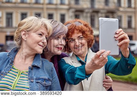 Three Smiling Older Caucasian Women Make Selfie In The Center Of A European City With A Tablet Compu