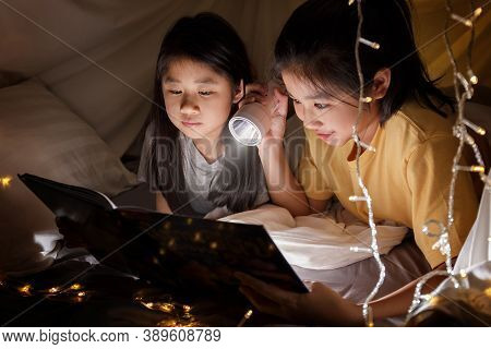 Family Concept. Elder Sister And Sister Reading Book With Flashlight Together In Children Tent Befor