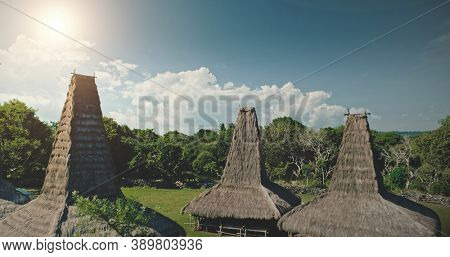 Sun light at high straw roof of traditional house in Kodi village, Indonesia. Aerial view of primitive Indonesian settlement admist green tropic forest. Amazing summer vacation at unique Asia landmark
