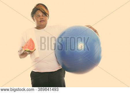 Studio Shot Of Fat Black African Woman Thinking While Holding Exercise Ball And Slice Of Watermelon 