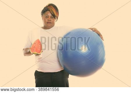 Studio Shot Of Fat Black African Woman Holding Exercise Ball And Slice Of Watermelon Ready For Gym