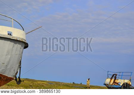Blue Sky. Old Rusty Abandoned Boats, White Ships Lie On Green Grassy Shore In The Light Of Setting S