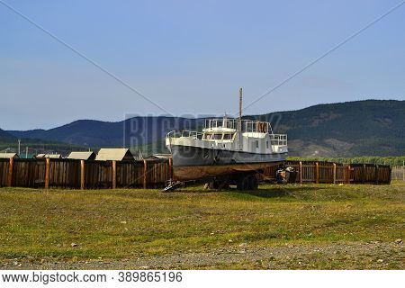 Old White Rusty Boat, Ship Stands By Wooden Fence In Village On Green Grassy Shore In Light Of Sunse
