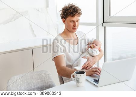 Attractive young father working on laptop computer while sitting at the table at home and holding his baby son
