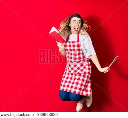 Young beautiful blonde cook woman wearing apron smiling happy. Jumping with smile on face holding recipe book and spoon over isolated red background