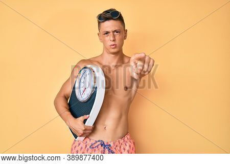 Young hispanic boy shirtless wearing swimwear and holding weighing machine pointing with finger to the camera and to you, confident gesture looking serious 