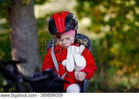 Little Toddler Girl With Security Helmet On Head Sitting In Bike Seat Of Her Mother Or Father Bicycl