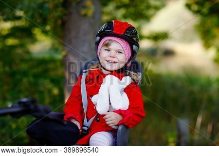 Little Toddler Girl With Security Helmet On Head Sitting In Bike Seat Of Her Mother Or Father Bicycl