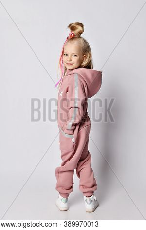 Smiling Blonde Kid Girl With Girlish Hairstyle And Colorful Braids In Stylish Pink Warm Jumpsuit Wit