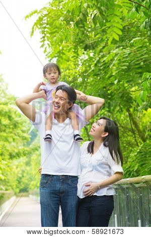 Happy Asian family outdoor. Father piggybacking his daughter walking in garden park with pregnant wife. Healthy lifestyle. 