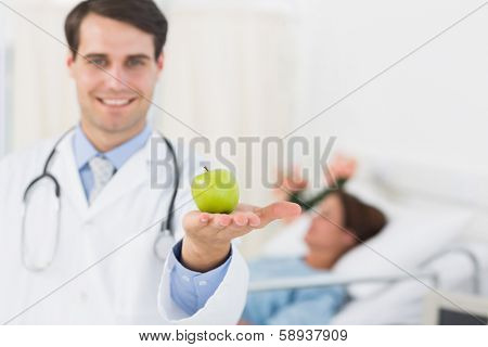 Portrait of a smiling doctor holding apple with patient in background at hospital
