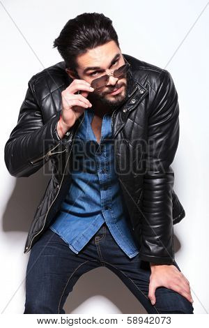 young man in leather jacket taking off his sunglasses and leaning against studio wall