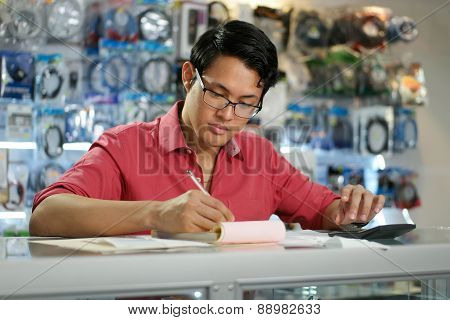 Chinese Man Working In Computer Shop Checking Bills And Taxes