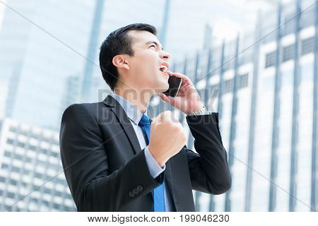 Winning businessman talking on mobile phone in the city clenching his fist feeling great and delighted - success and achievement concepts