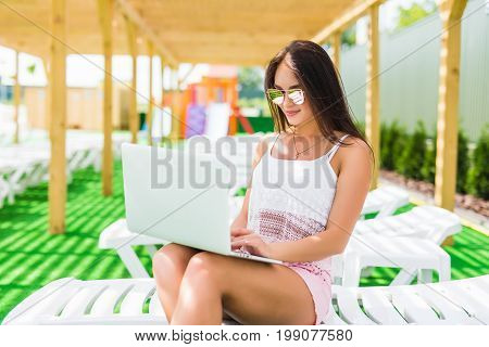 Working Even On Vacation. Attractive Young Woman In Sunglasses And Hat Is Lying On A Chaise-longue N