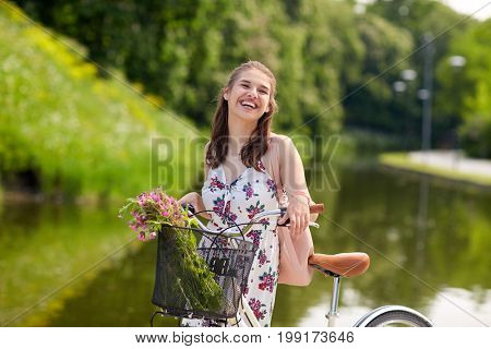 people, leisure and lifestyle - happy young hipster woman wearing summer dress riding fixie bicycle with wild flowers in basket at park
