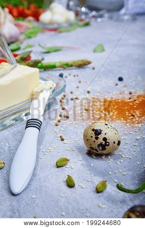 A close-up picture of breakfast ingredients. A glass container for butter next to a metal butter knife o a gray table background. Spices and quail egg. Green raw asparagus. Food preparation.