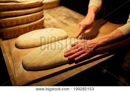 food, baking and people concept - chef or baker with knife cutting dough and cooking bread at bakery