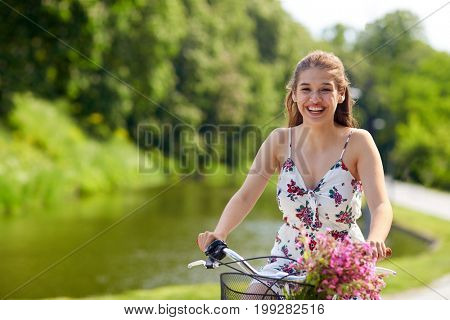 people, leisure and lifestyle - happy young hipster woman wearing summer dress riding fixie bicycle with wild flowers in basket at park