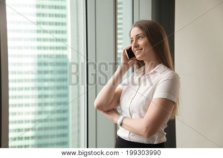 Smiling delighted businesswoman talking on cell while standing at office window, happy female manager making answering call in mobile roaming, lady having pleasant phone conversation with client