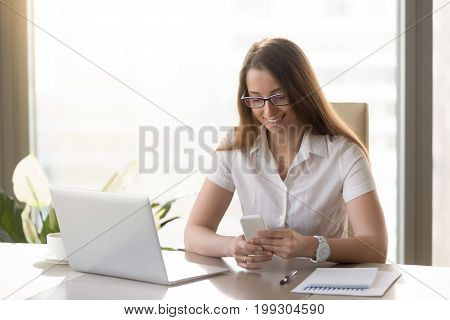 Beautiful smiling business lady using smartphone while sitting at desk, happy businesswoman holding new white cellphone, reading or texting message, dialing number to contact client, mobile banking
