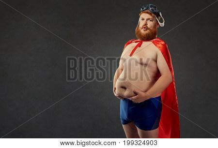 Thick fat funny man in swimming trunks in a superhero costume holds on to a large naked stomach.  The concept of weight loss.