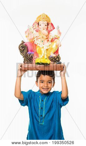 Portrait of cute little Indian boy holding a Ganesh idol or lord ganesha or ganapati murti /statue over his head, taking home on Ganesh Chaturthi, Isolated over white background