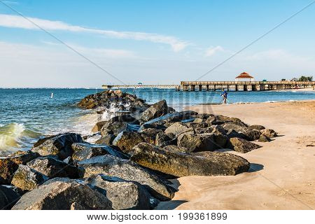 HAMPTON, VIRGINIA - JULY 9, 2017:  A man uses a beach metal detector near the rock jetty at Buckroe Beach in Hampton, Virginia, with the public viewing pier and fishing pier in the background.