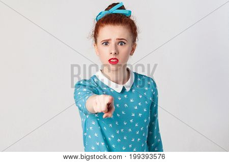 The seriosly redhead girl wearing blue dress opening mouths widely having surprised shocked looks pointing finger at camera. Isolated studio shot on gray background
