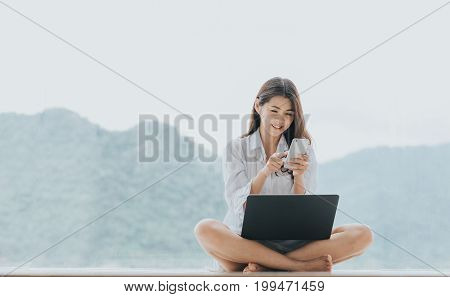 Happy Asian woman using smartphone mobile and laptop while sitting with mountain in background.