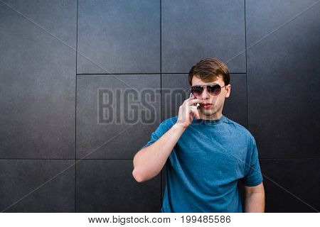 Handsome caucasian model dressed in trendy clothes and stylish sun glasses calling to best friend via cellular. Caucasian stylish man having mobile phone conversation while standing near copy space