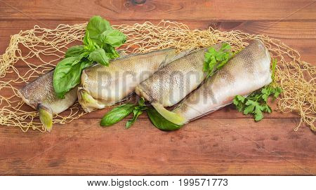 Several uncooked carcasses of the notothenia fish without od head and tail and twigs of basil and parsley on the fishing net on a dark wooden surface