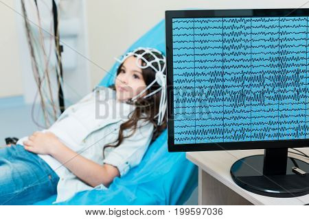 Informative examination. The focus being on a computer screen displaying recordings of brain waves of a charming little girl lying on an examination table