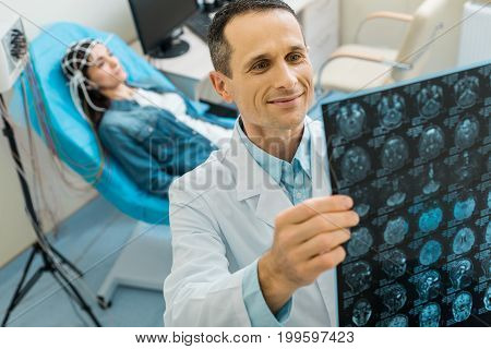 Content with results. Charming young doctor examining CT scan results of his female patient and looking pleased by them while the woman undergoing electroencephalography