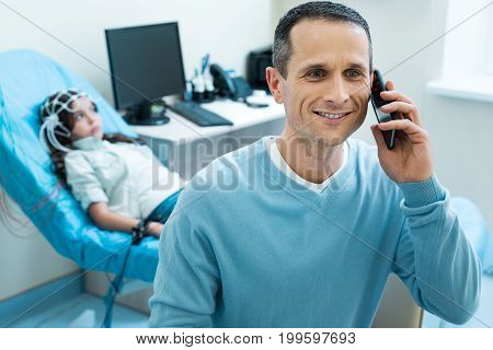 Great news. Cheerful handsome man talking on the phone, telling good news about his daughters condition while she undergoing electroencephalography in the background