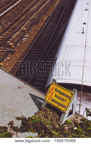 Yellow sign warning for high voltage contact wires above the rails, and white speed train in the background.