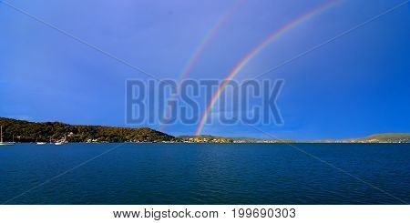 Spectacular colourful vivid rare Double Rainbow in blue sky with a tree studded horizon over water. Photographed at Lake Macquarie Central Coast New South Wales Australia.