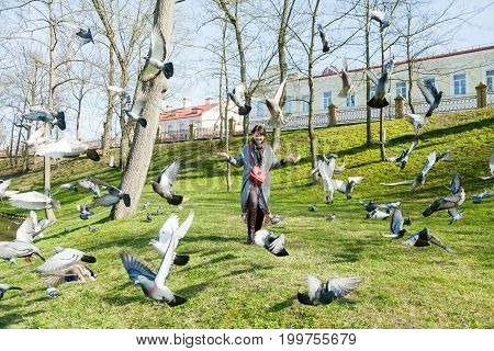 A girl in a gray coat runs through a flock of pigeons on the lawn on a sunny day