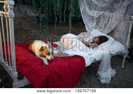 Young woman in lacy peignoir lies with dog on bed with bedding and baldachin near tree and sleeps. Concept of rest relaxation and serenity.