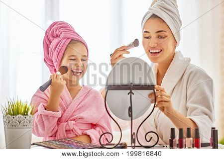 Happy loving family. Mother and daughter are doing make up and having fun sitting at dressing table at home. Mom and child girl are in bathrobes and with towels on their heads.