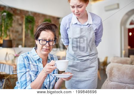 Middle-aged woman wearing checked shirt making wry face due to unsavory coffee, young red-haired waitress standing next to her with worried expression