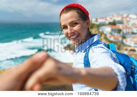 Follow me - happy young woman in a red hat and with a backpack behind her back pulling guy's hand. Hand in hand walking to the ocean coast near Azenhas do Mar Portugal. Concept of carefree modern life