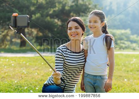 Gathering memories. Loving young mother squatting near her little daughter, hugging her and taking a selfie with a monopod