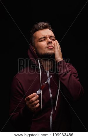 Portrait of a young male, suffering from pain, stress and drug addiction. A brunette guy in a red hoodie, holding a full syringe on a black background. Weakness, obsession, drug cravings concept.