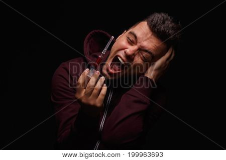 A narcomaniac guy in a dark hoodie, holding a syringe and screaming on a black background. A young man fighting with drug cravings. Cocaine, meth, cannabis, LSD, ecstasy, morphine addiction concept.
