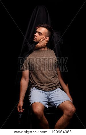 Dead narcomaniac on a chair holding a syringe with narcotics on a black background. Death in black clothing holding a head of an addicted to morphine or LSD young man. Deadly addictive drugs concept.